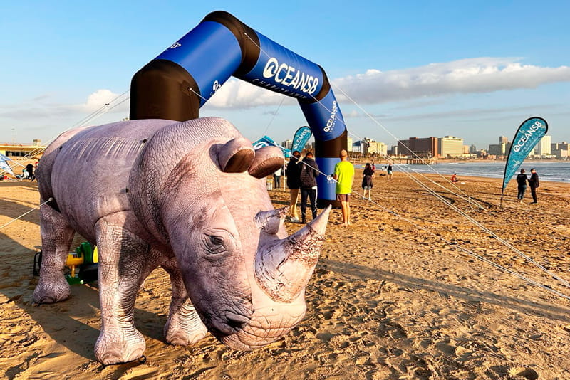 Aufblasbares Nashorn am Strand Aufblasbares Nashorn 'Nashorn Blow Up' steht am Strand unter einem Bogen mit der Aufschrift 'OCEANSB'.