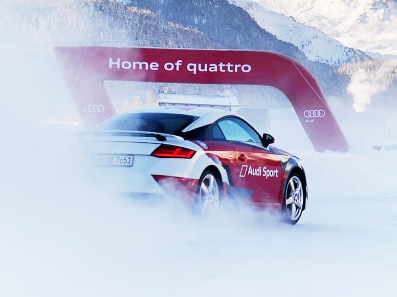 Audi TT auf verschneiter Strecke mit Werbebanner Audi TT Sportwagen auf verschneiter Strecke, hinter ihm ein aufblasbares Werbebanner mit der Aufschrift 'Home of quattro'.