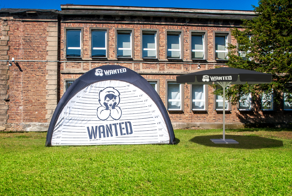 Aufblasbares Zelt und Schirm Zelt mit 'WANTED'-Aufdruck und Sonnenschirm auf einer Wiese vor einem Backsteingebäude.