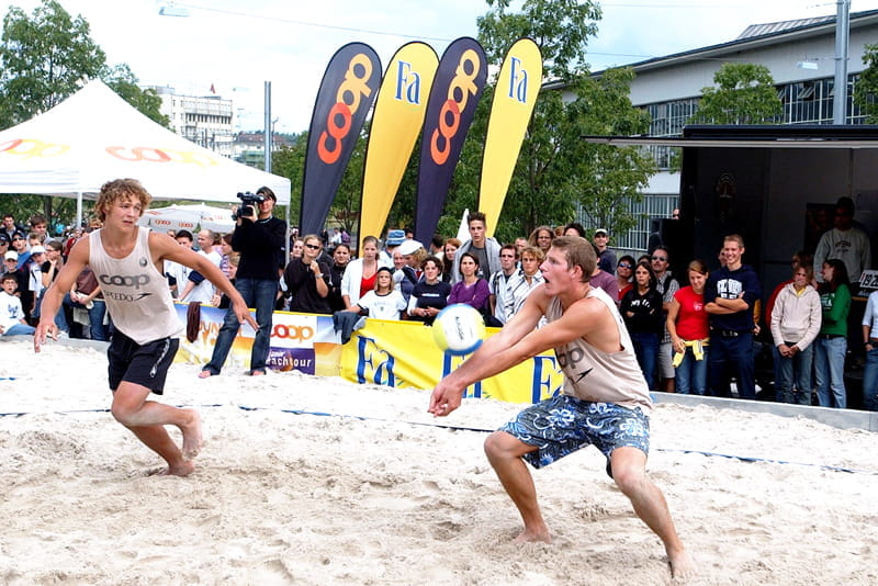 Beachvolleyball-Spiel mit Zuschauern und Beachflags Zwei Männer spielen Beachvolleyball im Sand, umgeben von Zuschauern und Beachflags von Coop und Fa.