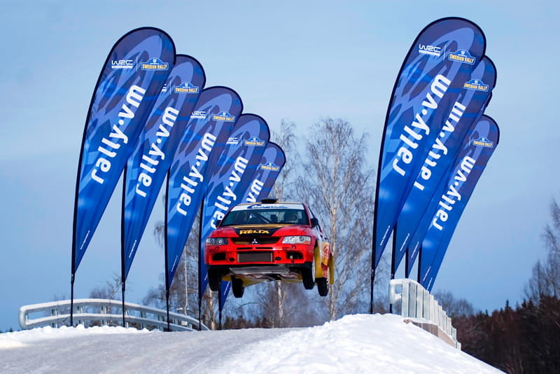 Rallye-Auto bei Winter Rallye mit Beachflags Ein Rallye-Auto springt über eine Schnee- und Eisfläche, umgeben von Beachflags mit dem Schriftzug 'rally.vum'.