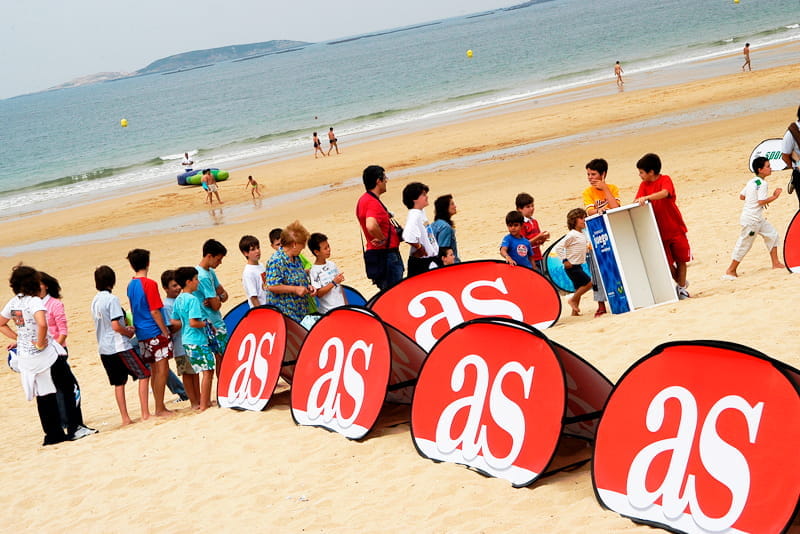 Werbebande Quickboard Strand am Strand Werbebande Quickboard Strand mit dem Logo 'as' am Strand, umgeben von Kindern und einem Kiosk.