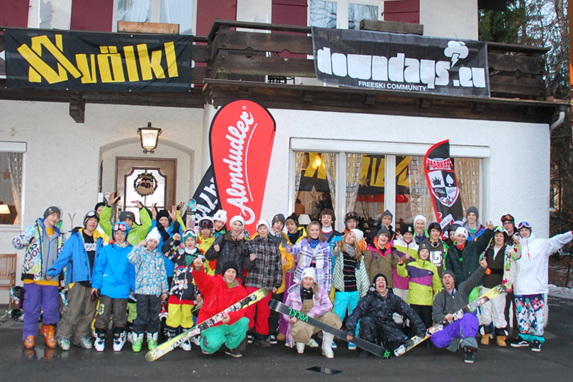 Gruppe von Skifahrern posiert vor einer roten Beachflag und einer Skihütte mit Werbebanner