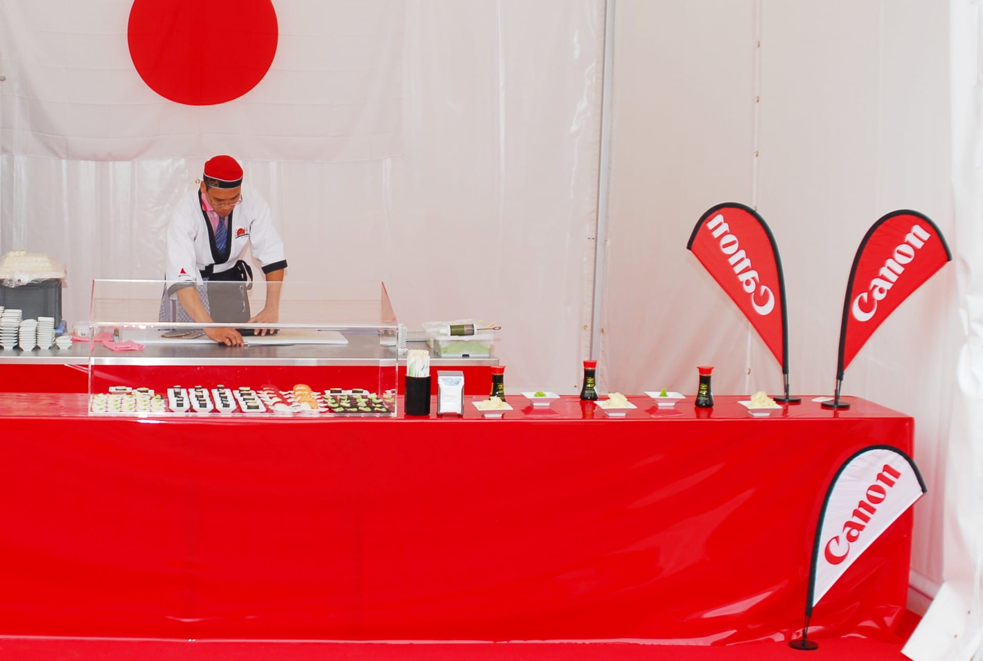 Sushi-Stand mit Canon-Flaggen Ein Sushi-Stand mit rotem Tisch und Canon-Flaggen. Ein Koch bereitet Sushi vor.