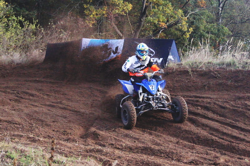 Falcon Motorsport Quad Fahrer Mobile Werbebande Clickboard Ein Quadfahrer in blauer Rennkleidung fährt auf einem unebenen, erdigen Gelände neben einer mobilen Werbebande von ENTDECKER.