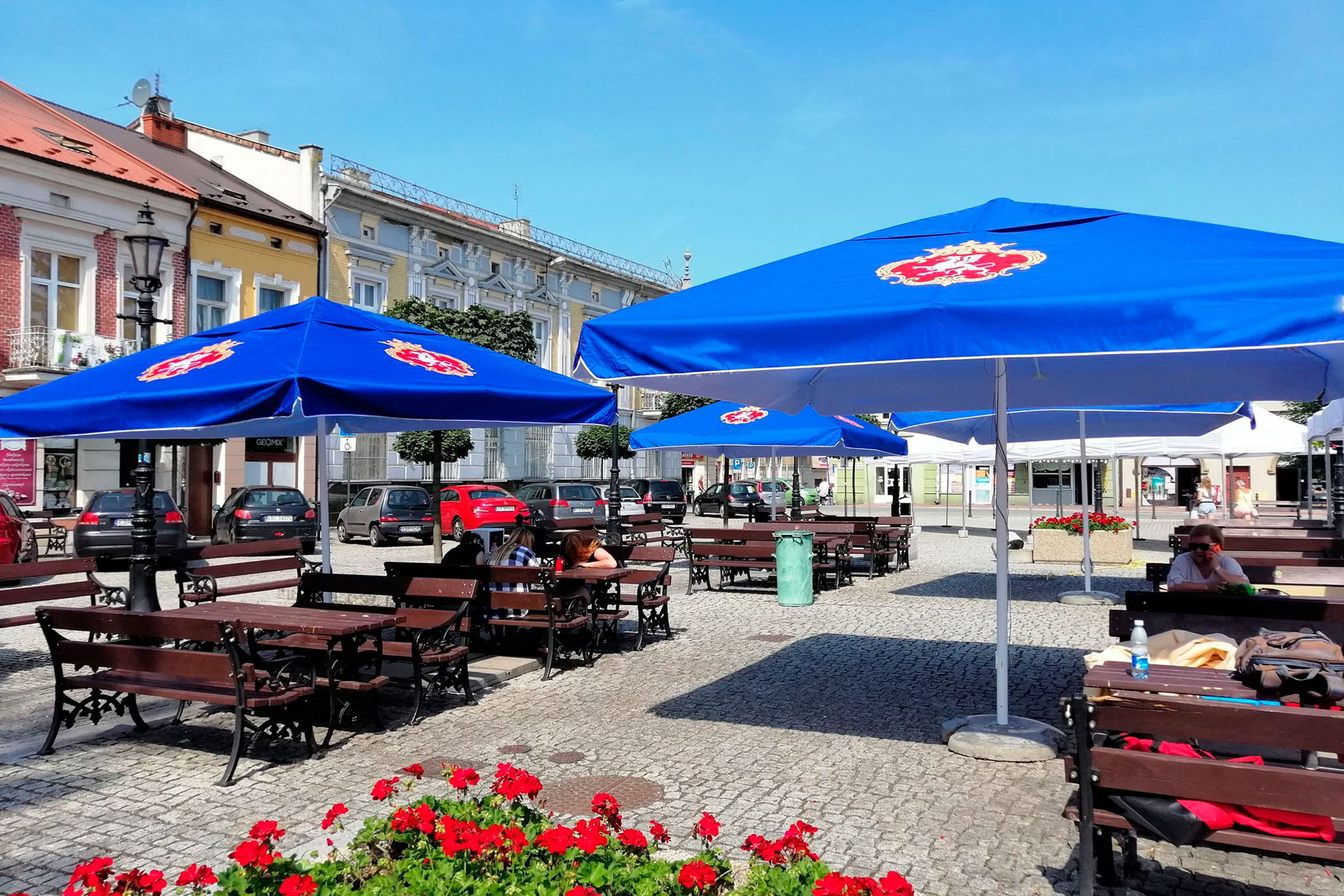 Sonnenschirm Expert Biergarten Biergarten mit blauen Sonnenschirmen des Produkts 'Sonnenschirm Expert Biergarten', Tischen und Stühlen auf einem Platz.